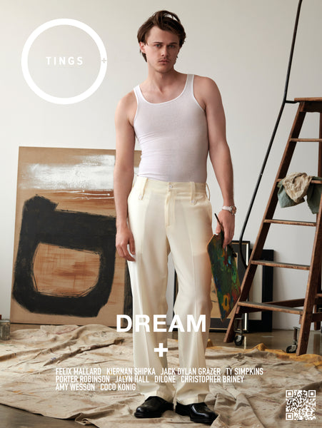 American Actor Christopher Briney stands ini an artist's studio holding a painter's palette and paint brush wearing white linen Prada pants, a white tank top, and a watch by Omega