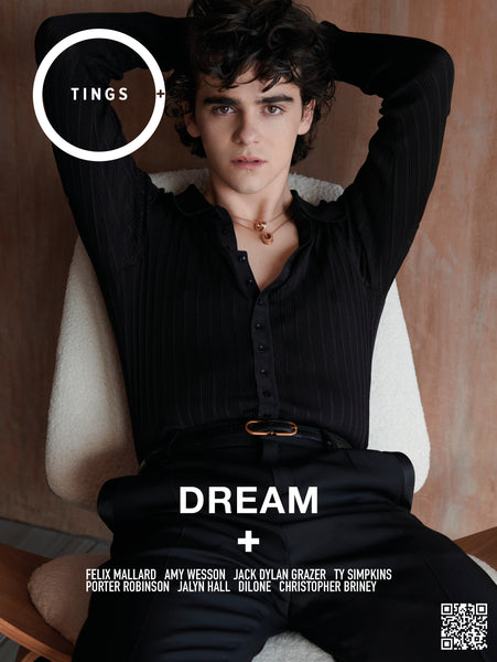 American Actor Jack Dylan Grazer leans back with his arms behind his head via mid century modern chair wearing a black button down knit shirt and black pants from Saint Laurent and two gold necklaces from Bulgari 